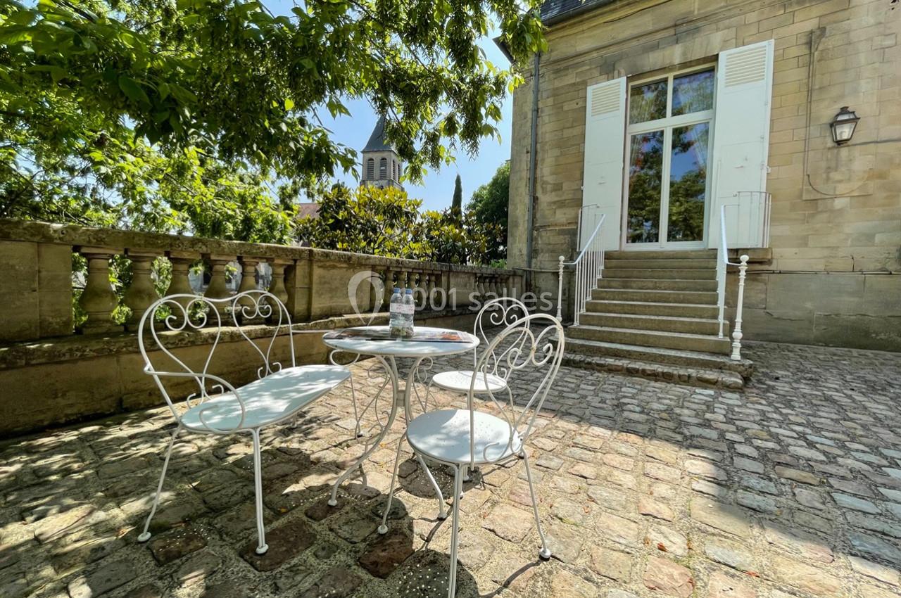 Terrasse pavée avec table et chaises blanches en fer forgé, devant une maison en pierre aux volets ouverts.