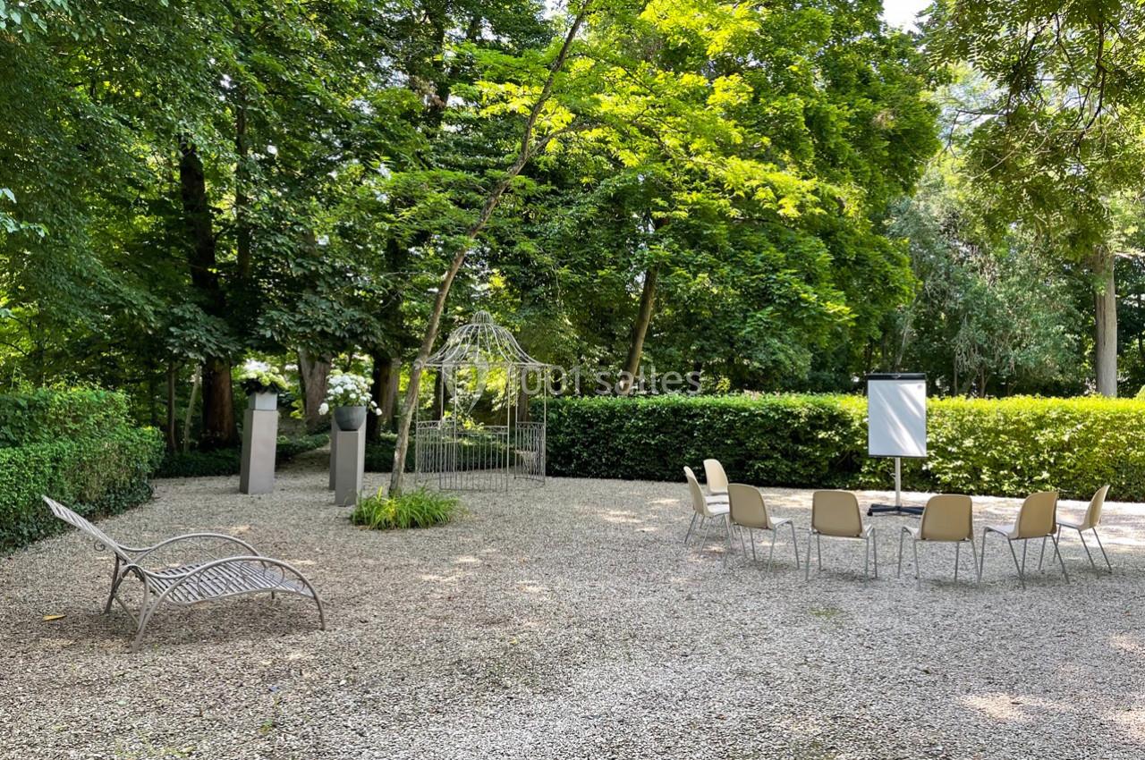 Espace extérieur avec des chaises disposées en cercle, un chevalet blanc et une tonnelle entourée de verdure.