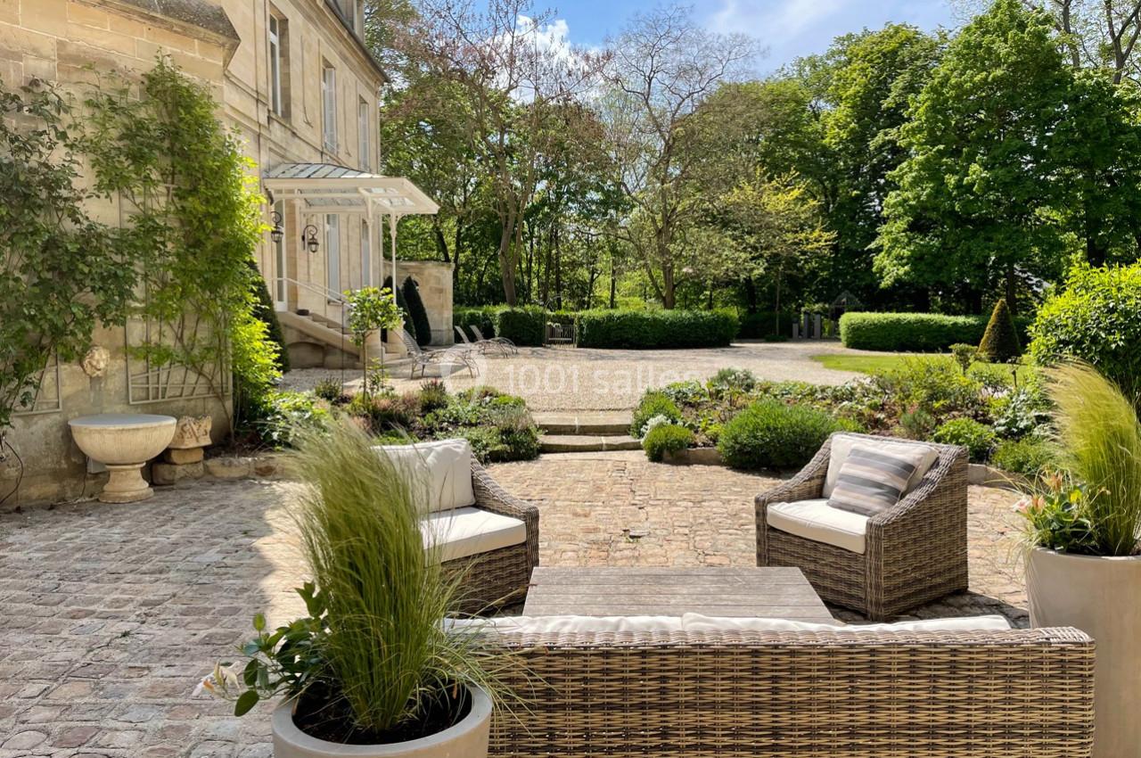 Terrasse pavée avec mobilier en rotin, plantes en pot et vue sur un jardin arboré devant une maison en pierre.