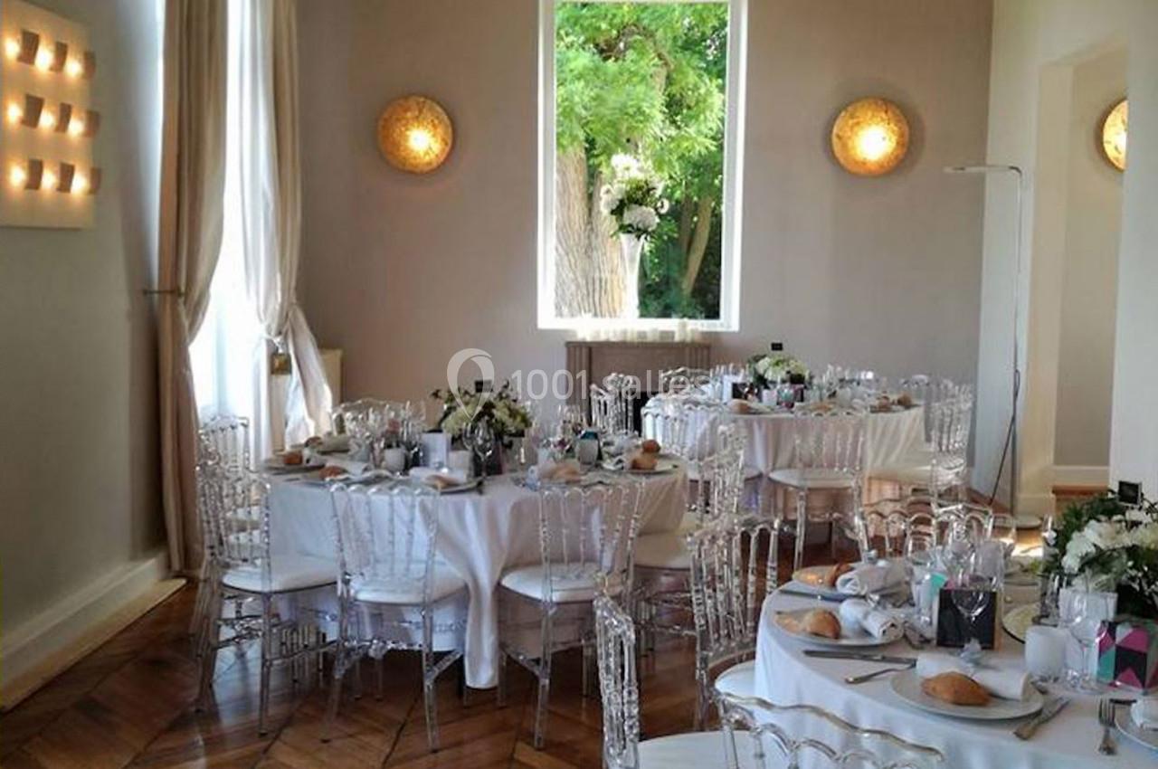 Salle de réception lumineuse avec tables rondes dressées, chaises transparentes et grande fenêtre donnant sur un jardin…