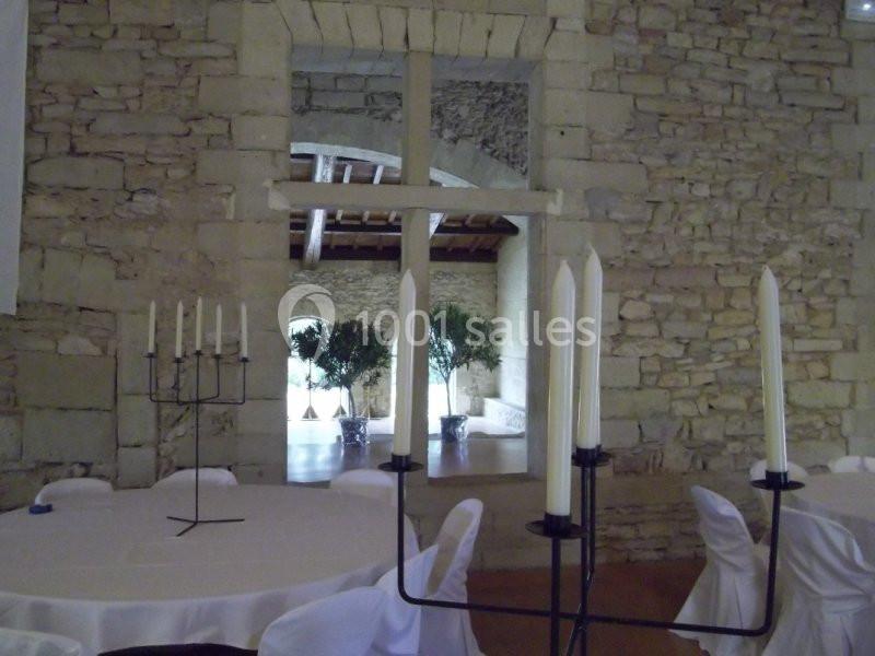 Salle en pierre avec des tables rondes dressées, des chandeliers et une grande croix en bois au centre.