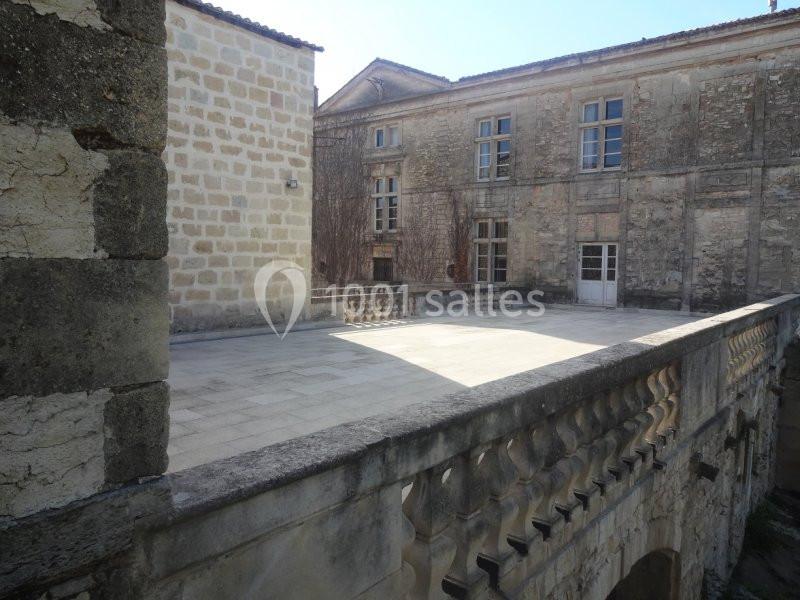 Cour pavée entourée de bâtiments anciens en pierre, avec balustrade en premier plan et ciel dégagé.