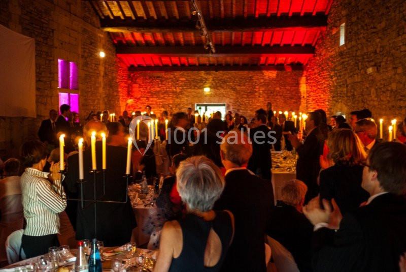 Salle en pierre éclairée par des bougies, remplie d'invités debout lors d'un événement festif.
