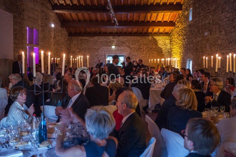 Salle en pierre éclairée par des bougies, avec des invités assis à des tables lors d'un événement formel.