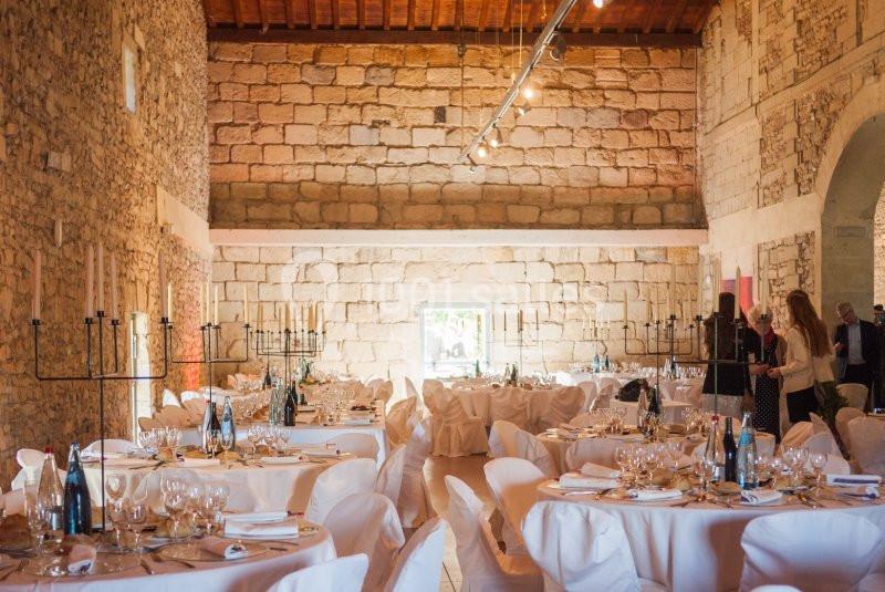 Salle en pierre avec tables rondes dressées pour un événement, décorée de chandeliers et éclairée par des lumières…