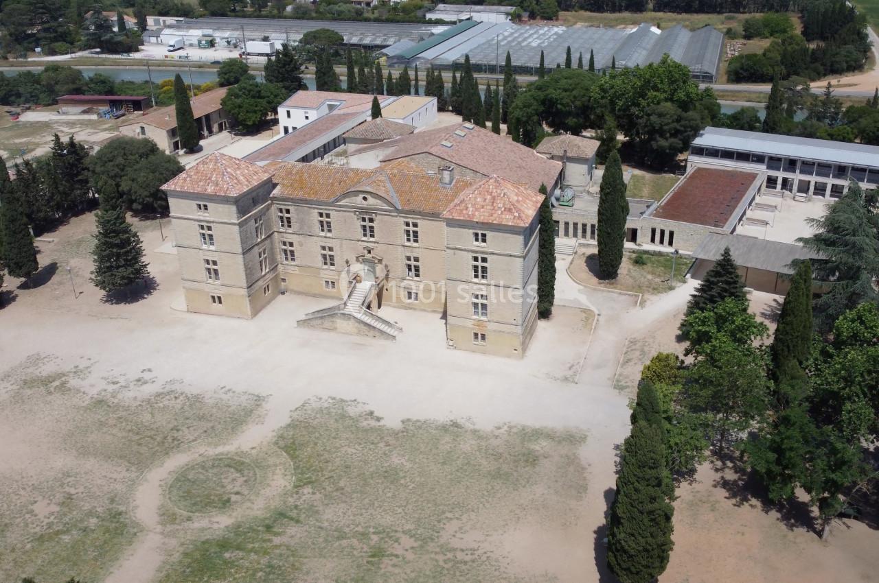 Vue aérienne d'un grand bâtiment historique entouré d'arbres, avec des serres visibles en arrière-plan.