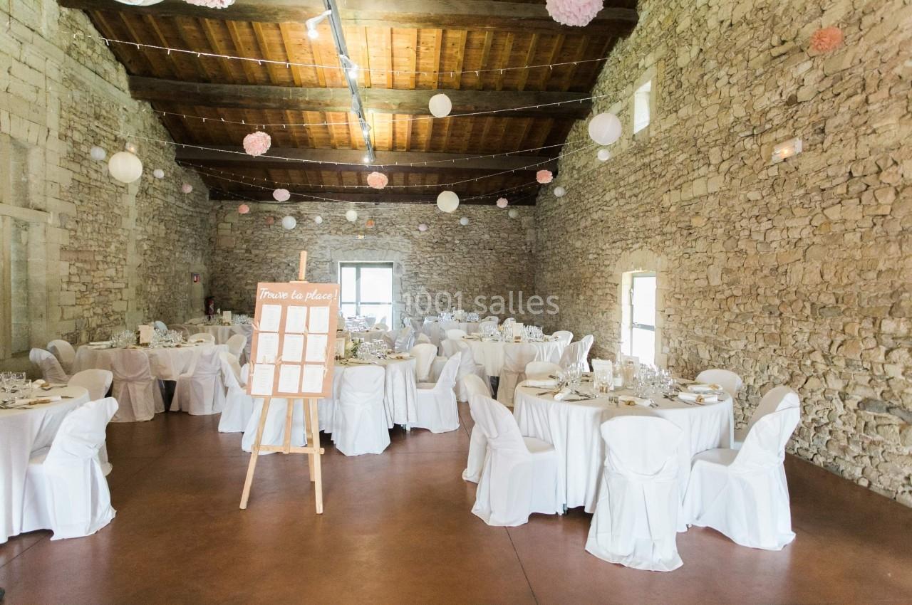 Salle en pierre décorée pour un événement, avec des tables rondes dressées et un plan de table sur un chevalet.