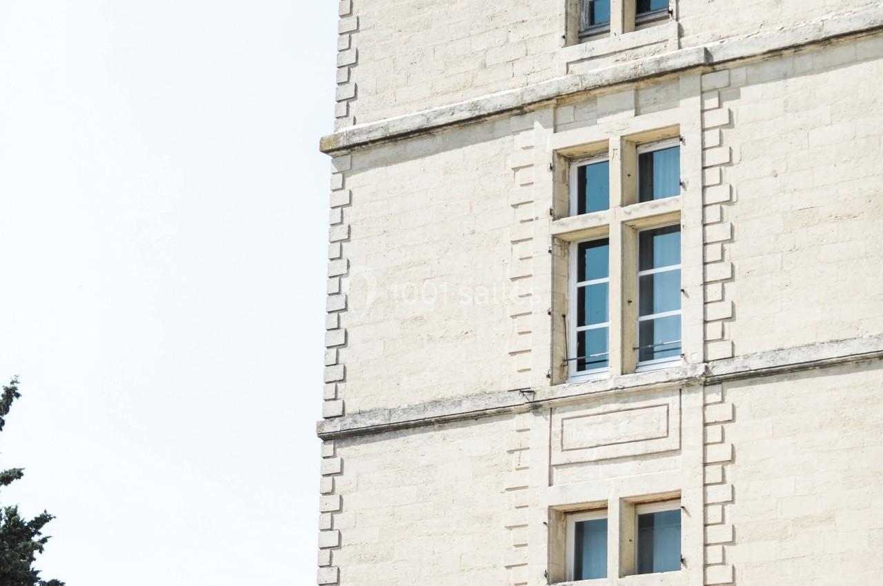 Façade d'un bâtiment en pierre claire avec des fenêtres à croisillons, entouré d'arbres sous un ciel dégagé.