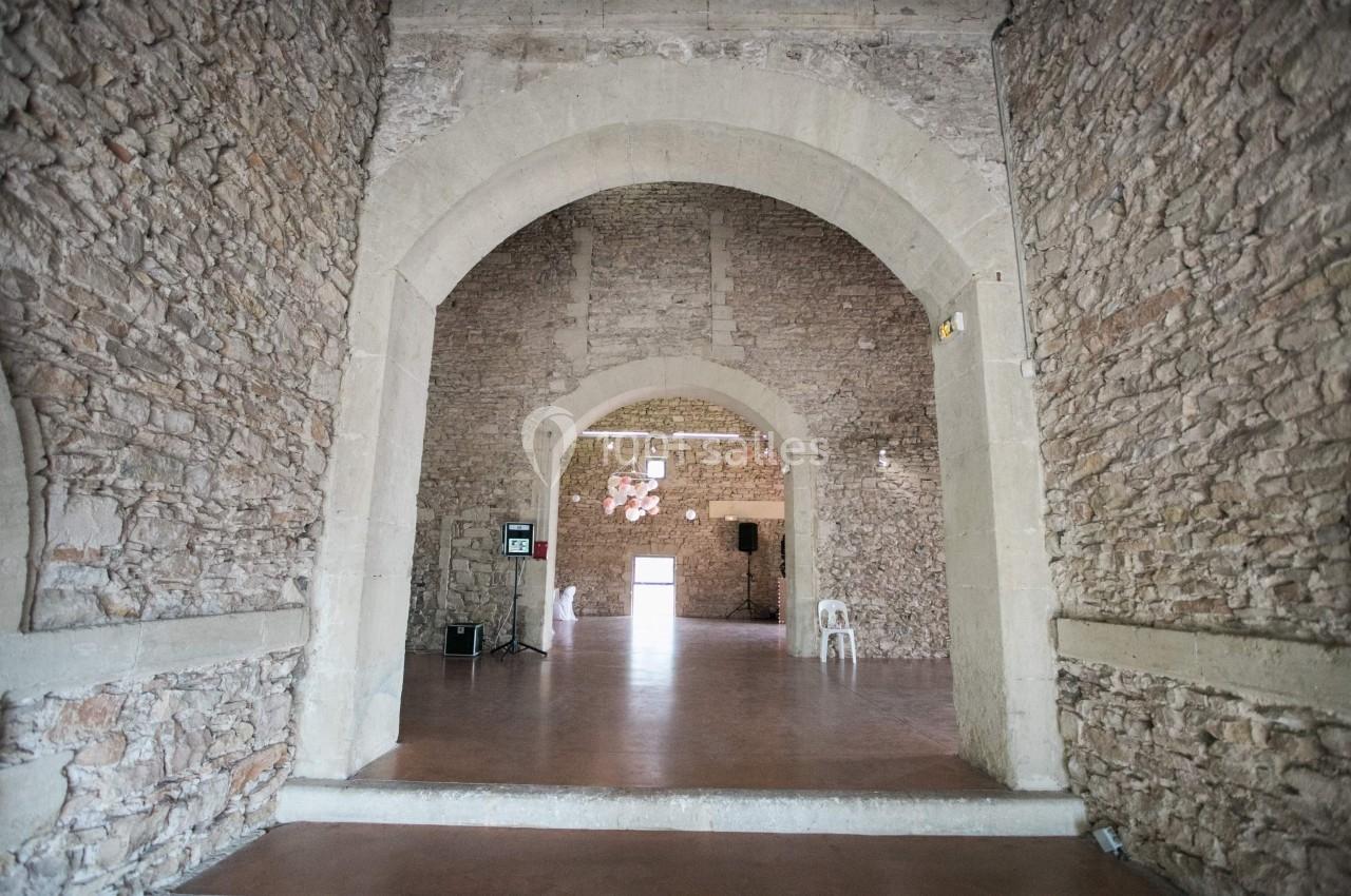 Vue intérieure d'une salle en pierre avec de grandes arches, un sol en béton et quelques chaises dispersées.
