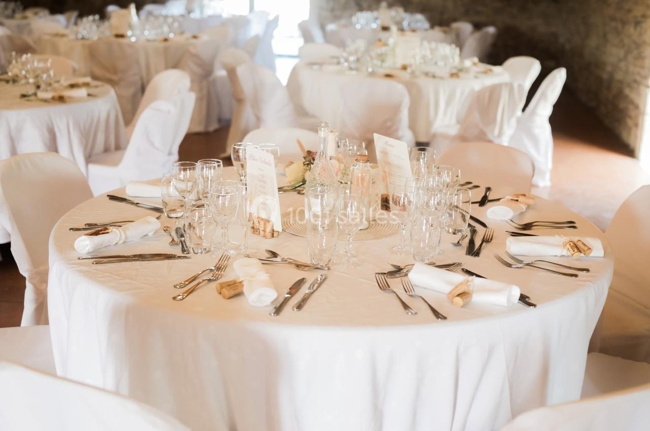 Table ronde dressée pour un événement, avec nappes blanches, couverts, verres et menus au centre.