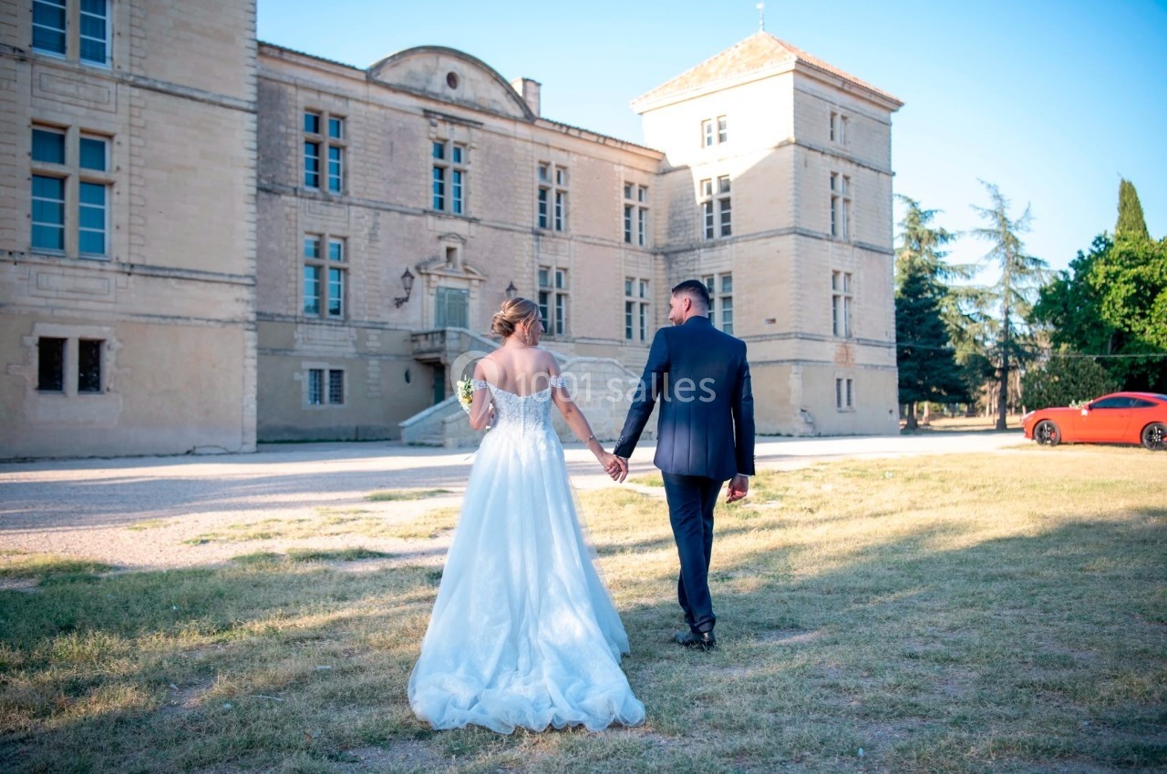 Un couple en tenue de mariage marche main dans la main devant un bâtiment historique en pierre par une journée ensoleillée.