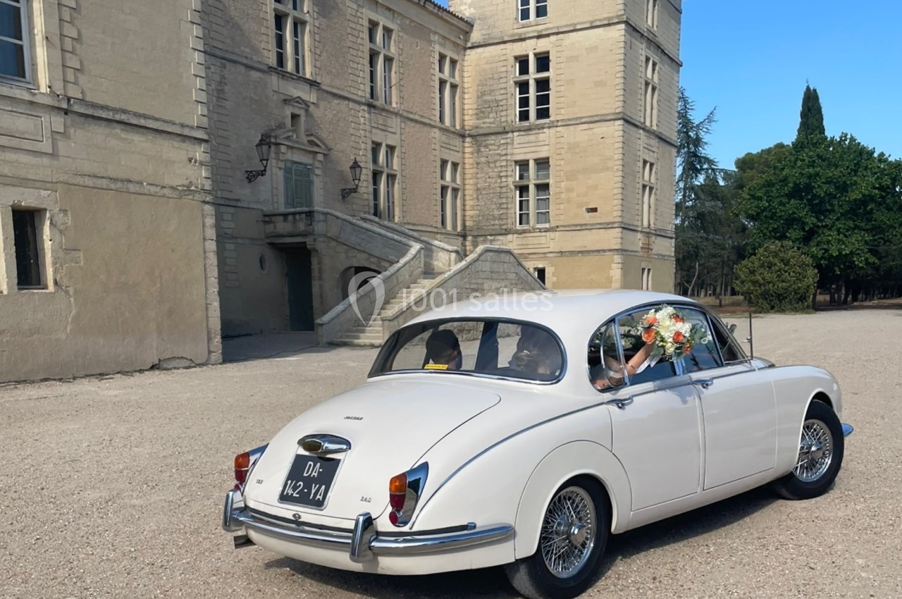 Voiture ancienne blanche devant un bâtiment historique en pierre, avec un bouquet de fleurs à l'arrière.