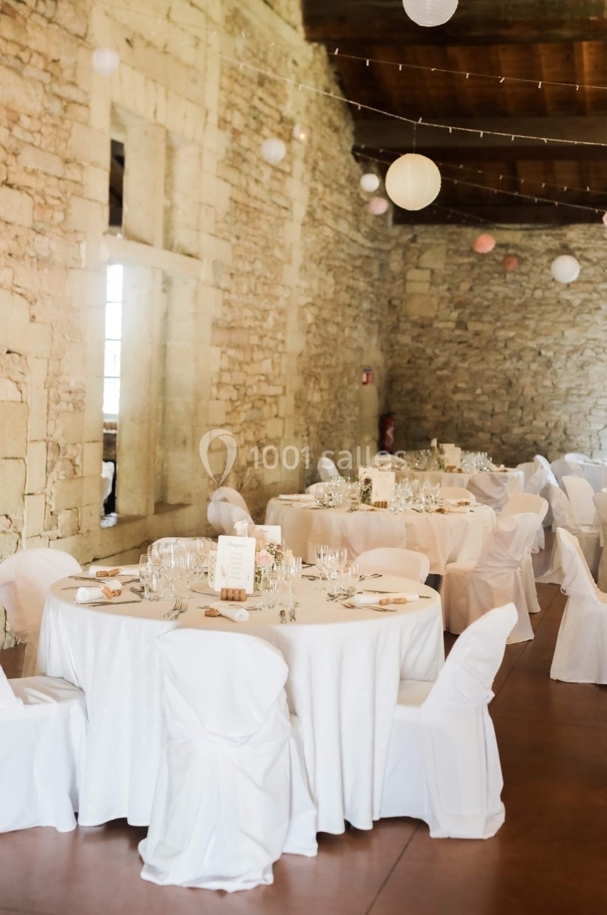 Salle en pierre décorée pour un événement, avec des tables rondes dressées et des chaises couvertes de housses blanches.