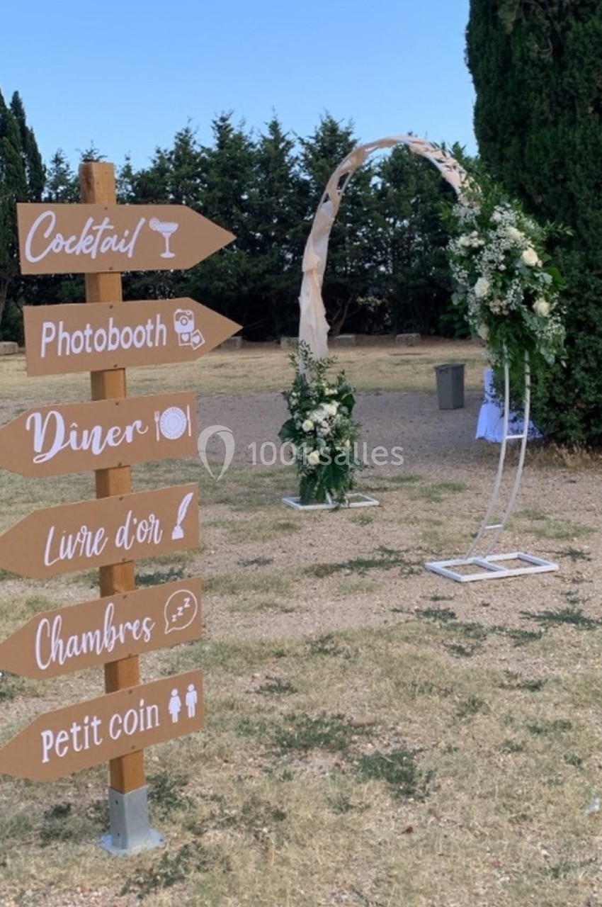 Panneau directionnel en bois indiquant les zones d'un mariage, avec une arche fleurie en arrière-plan.