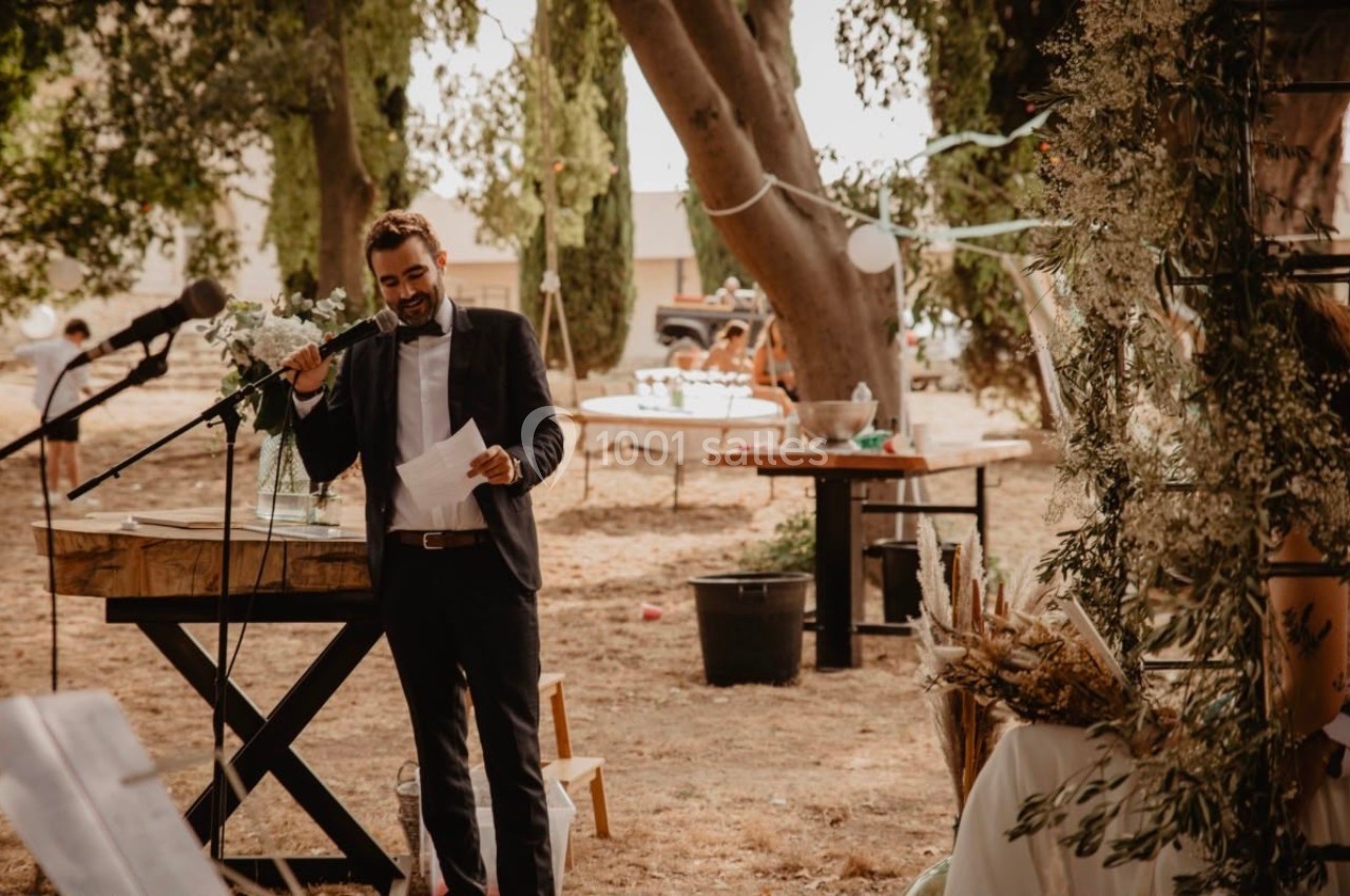 Un homme en costume parle au micro lors d'un événement en extérieur, entouré de décorations et d'arbres.
