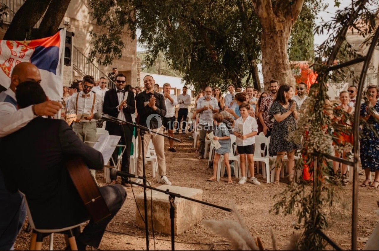 Un groupe de personnes assiste à une cérémonie en plein air sous des arbres, avec un musicien jouant de la guitare.