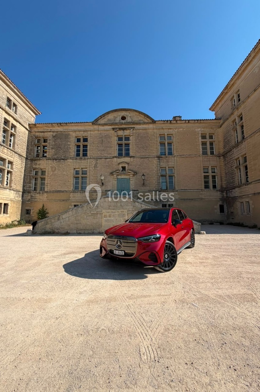 Voiture rouge garée devant un bâtiment ancien en pierre sous un ciel bleu clair.