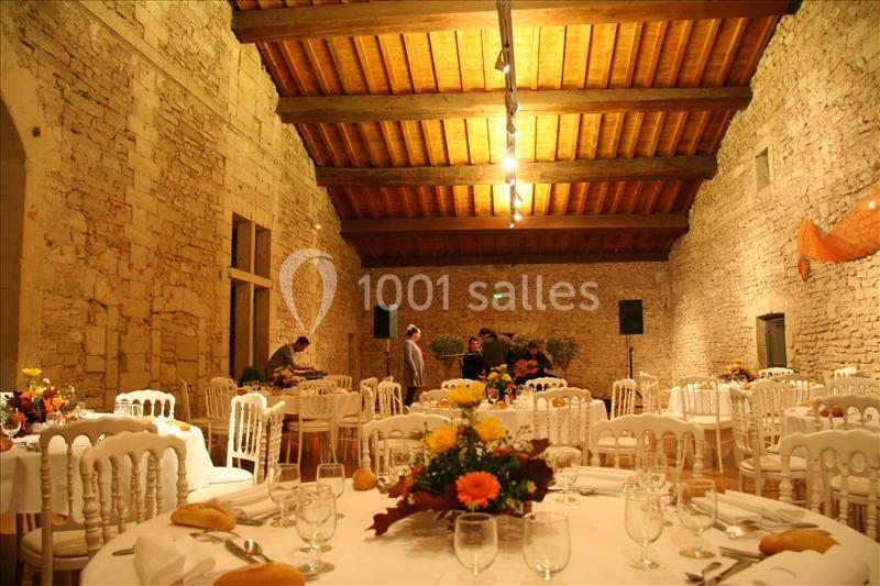 Salle en pierre avec plafond en bois, tables rondes décorées de fleurs et couverts pour un événement festif.