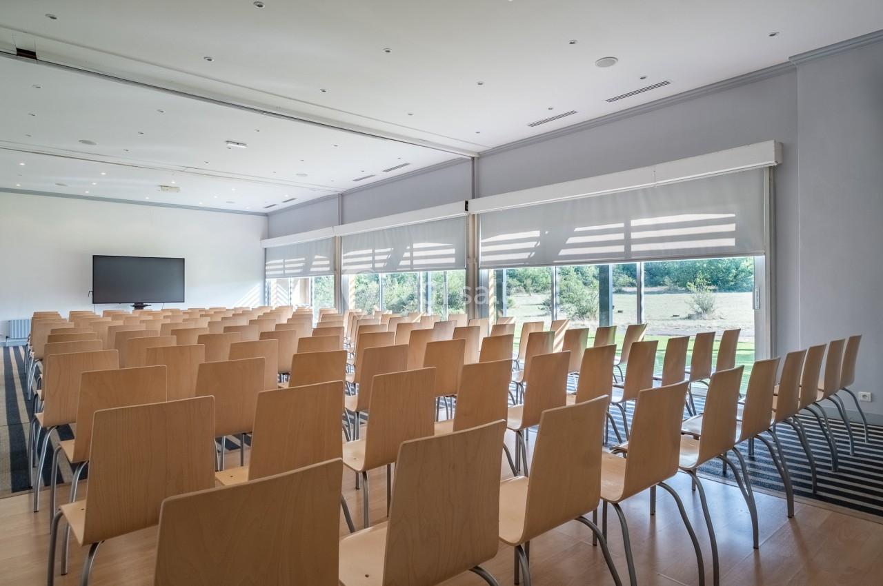 Salle de conférence lumineuse avec des rangées de chaises en bois, un écran et de grandes fenêtres donnant sur un espace…