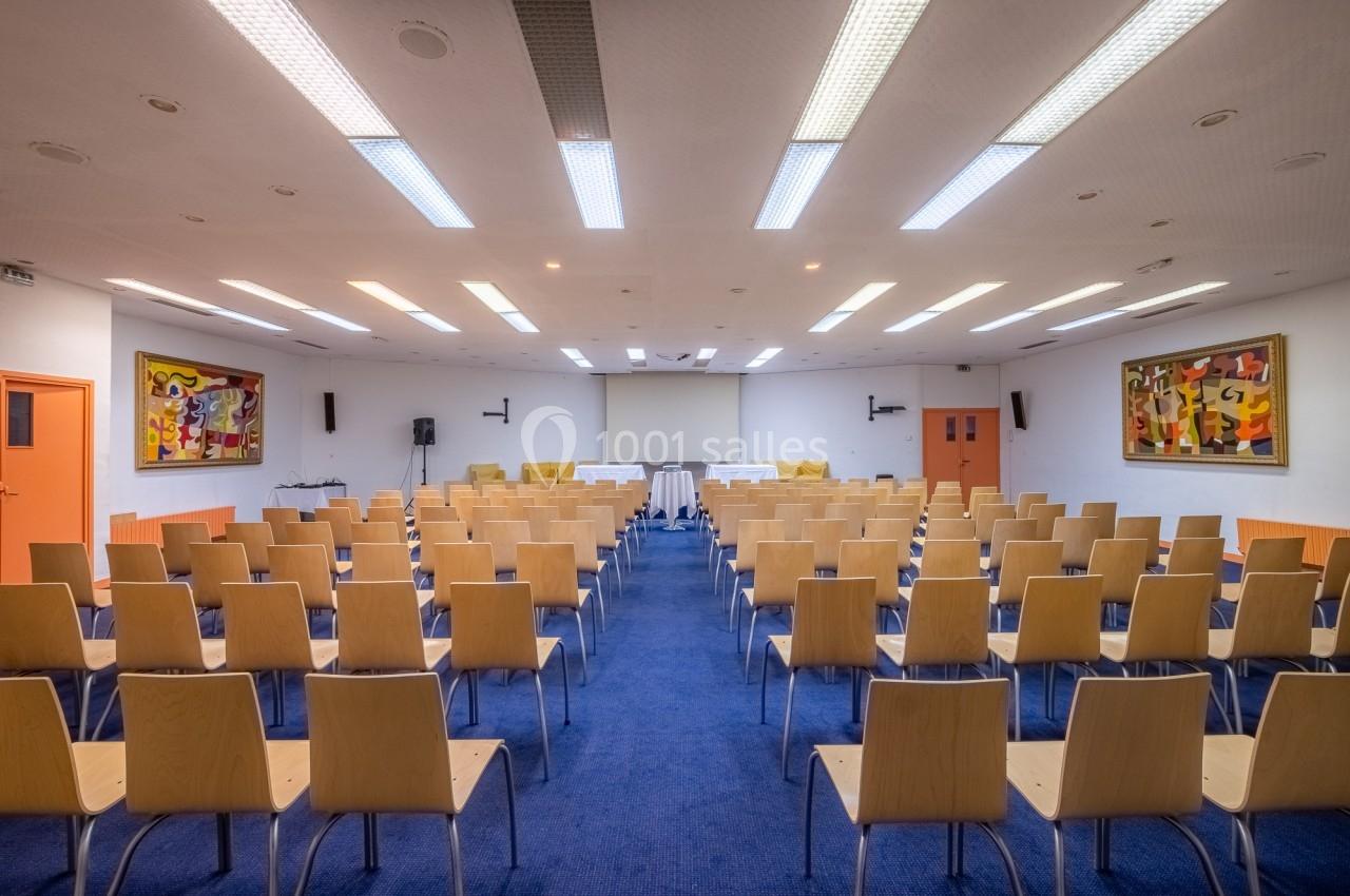 Salle de conférence avec rangées de chaises en bois, écran de projection et tableaux colorés aux murs.