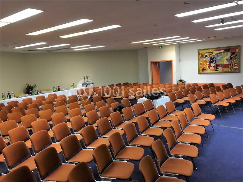Salle de conférence vide avec des rangées de chaises orange, un buffet à l'arrière et un tableau coloré au mur.