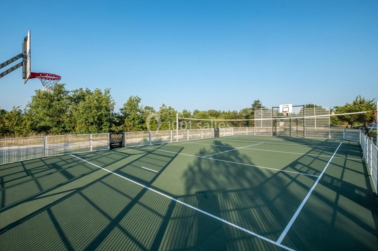 Terrain multisport extérieur avec panier de basket, filet de volley et clôture, entouré de végétation sous un ciel dégagé.