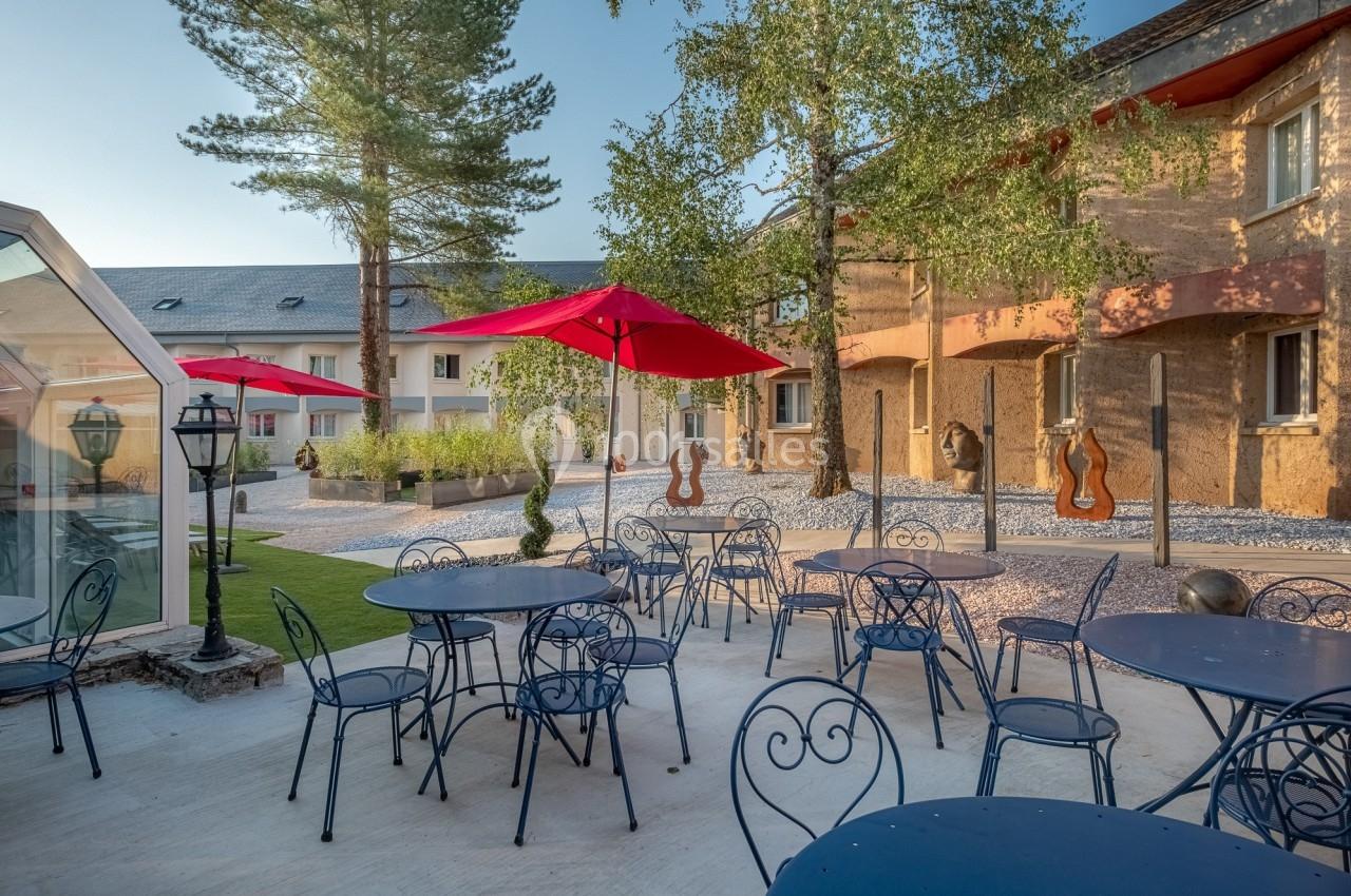 Terrasse extérieure avec tables et chaises en métal, parasols rouges et arbres, entourée de bâtiments en pierre.