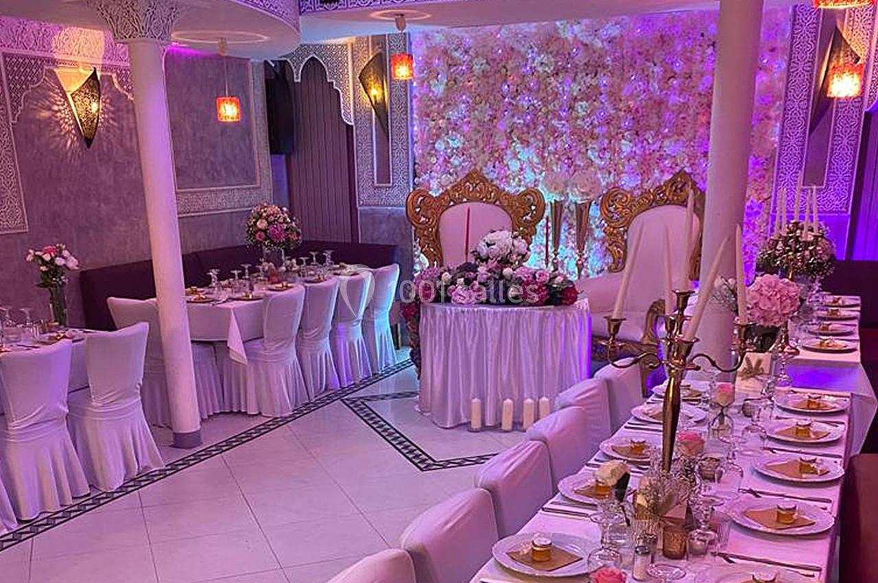 Salle de réception décorée avec des tables dressées, des chaises blanches et un fond floral illuminé.