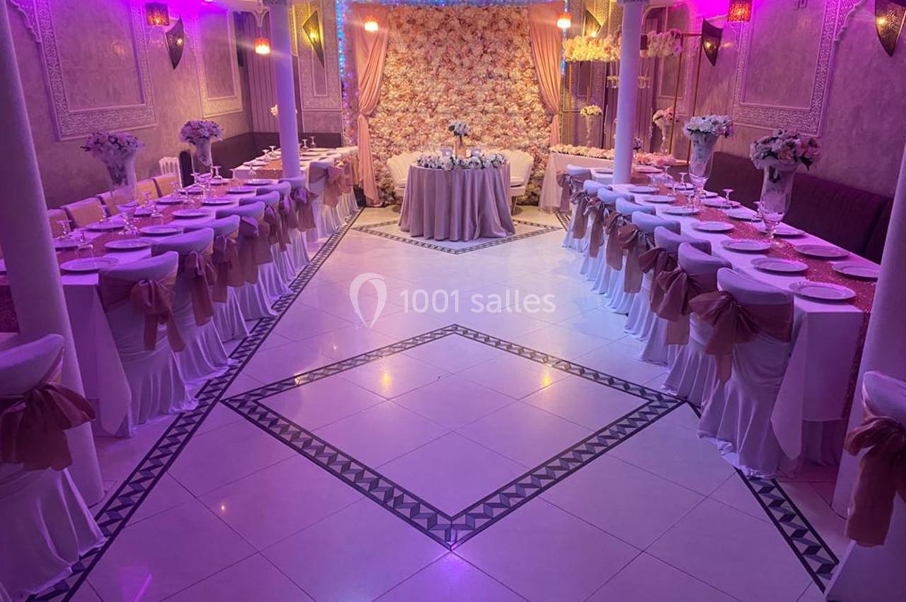 Salle de réception décorée avec des tables alignées, nappes blanches, chaises ornées de rubans et un mur floral en arrière…