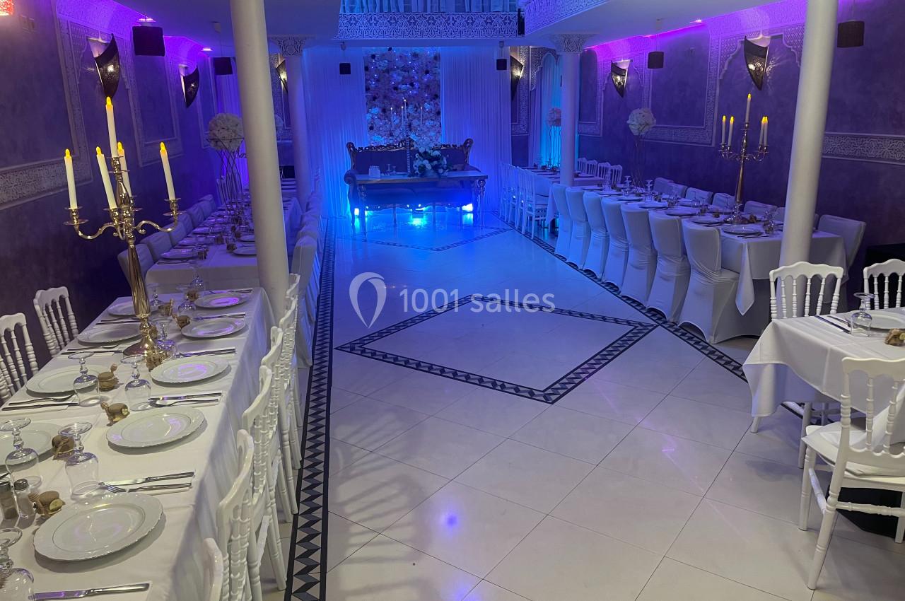 Salle de réception décorée avec des tables dressées, des chaises blanches et un éclairage violet.