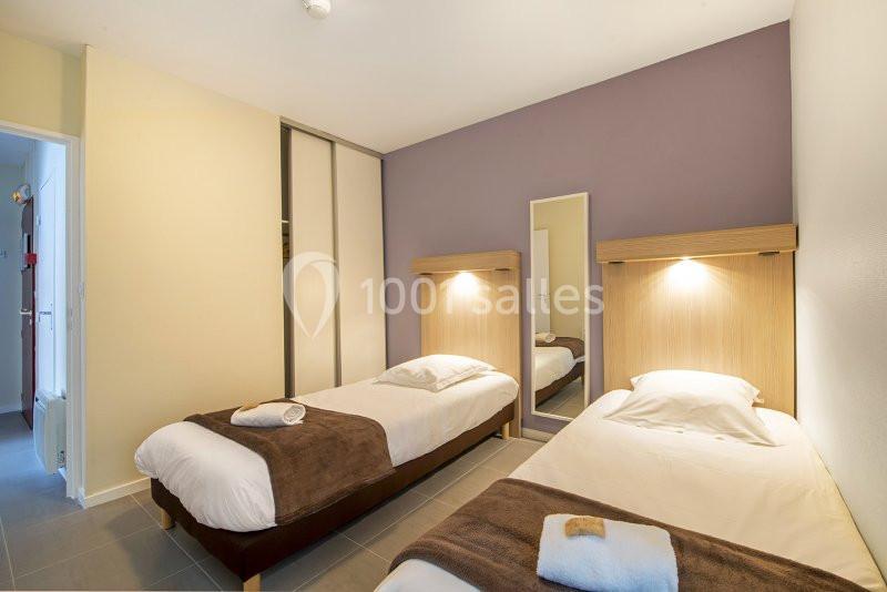 Deux lits simples avec couvertures brunes dans une chambre lumineuse, mur violet, armoire et miroir.