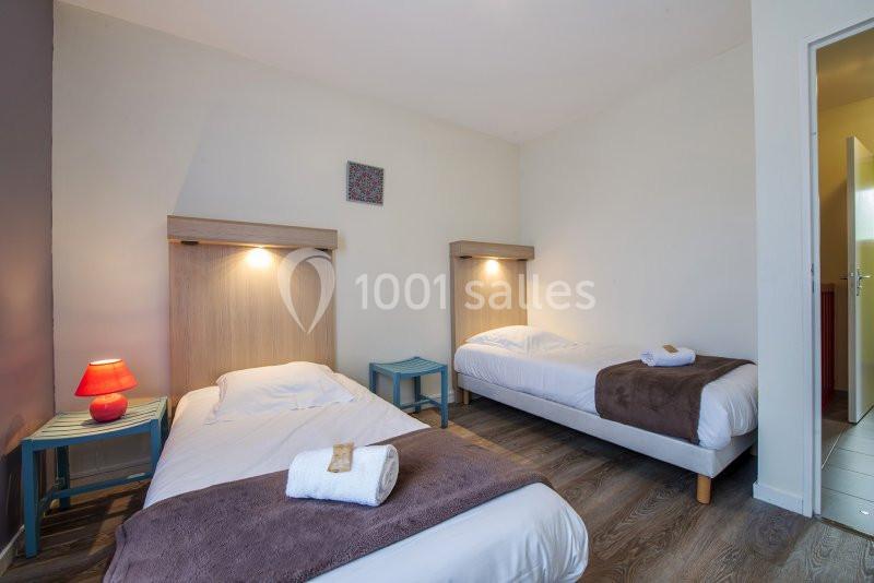 Deux lits simples avec linge de lit blanc et couvertures brunes dans une chambre lumineuse au sol en parquet.