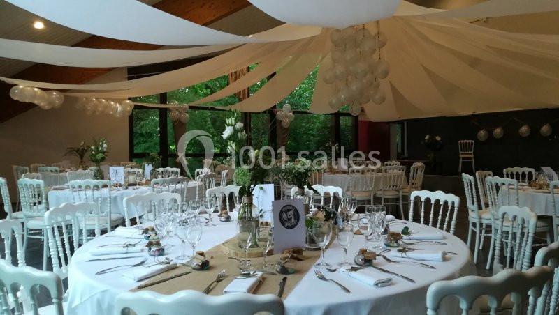 Salle décorée pour un événement avec tables rondes dressées, nappes blanches, chaises assorties et guirlandes suspendues.