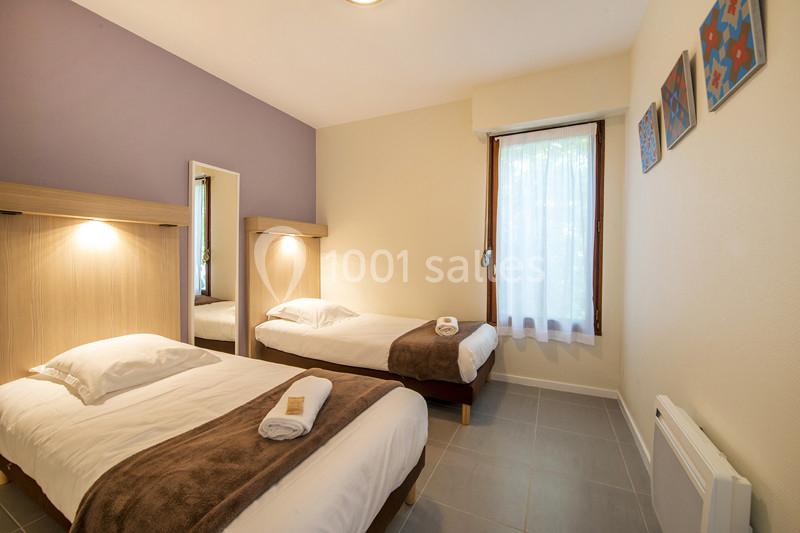 Deux lits simples avec couvertures marron dans une chambre lumineuse, décorée de tableaux et d'un mur violet.