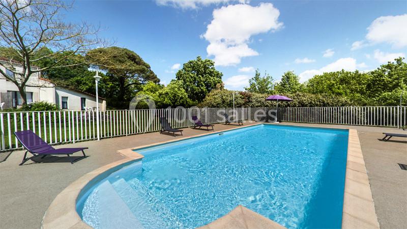 Piscine extérieure entourée de chaises longues et d'arbres, avec ciel bleu et quelques nuages en arrière-plan.