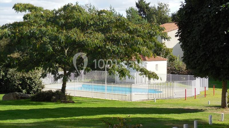 Piscine extérieure entourée d'une clôture blanche, située dans un jardin avec pelouse, arbres et bâtiments en arrière-plan.