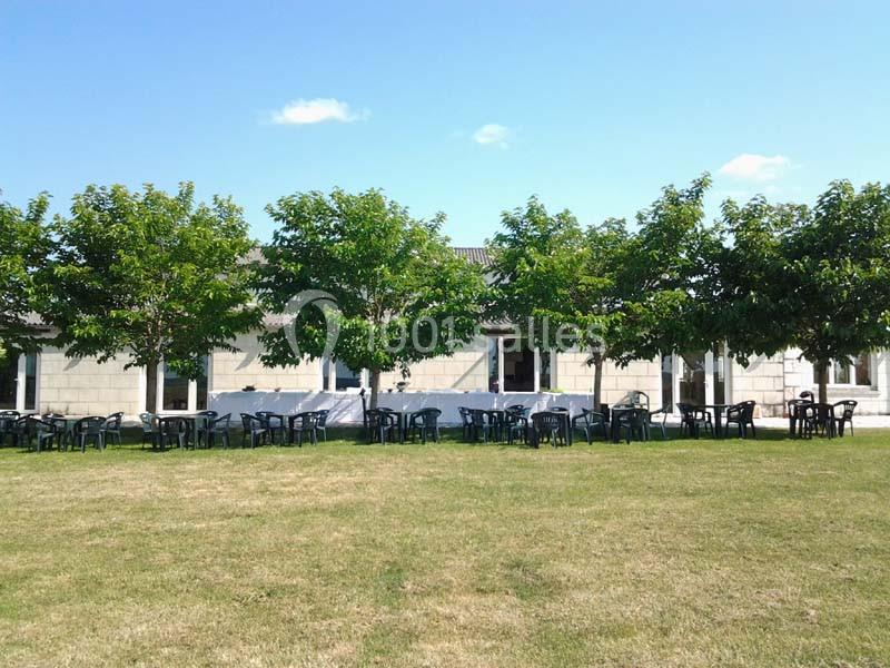 Des tables et chaises en plastique noir sont disposées sous des arbres devant un bâtiment en pierre par une journée…