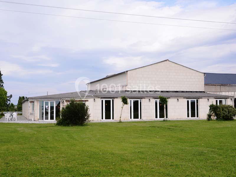 Bâtiment en pierre clair avec de grandes baies vitrées, entouré d'une pelouse et d'arbustes sous un ciel nuageux.