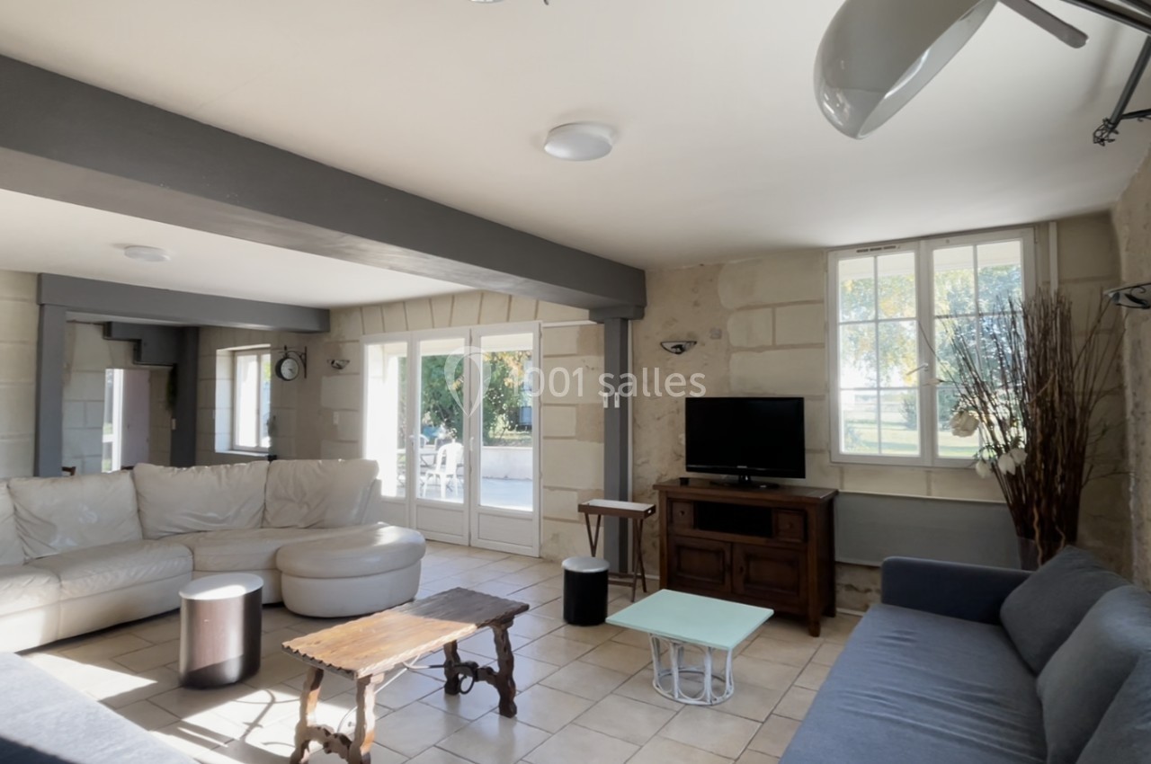 Salon lumineux avec canapé d'angle, table basse en bois, télévision sur meuble et grandes fenêtres donnant sur un jardin.