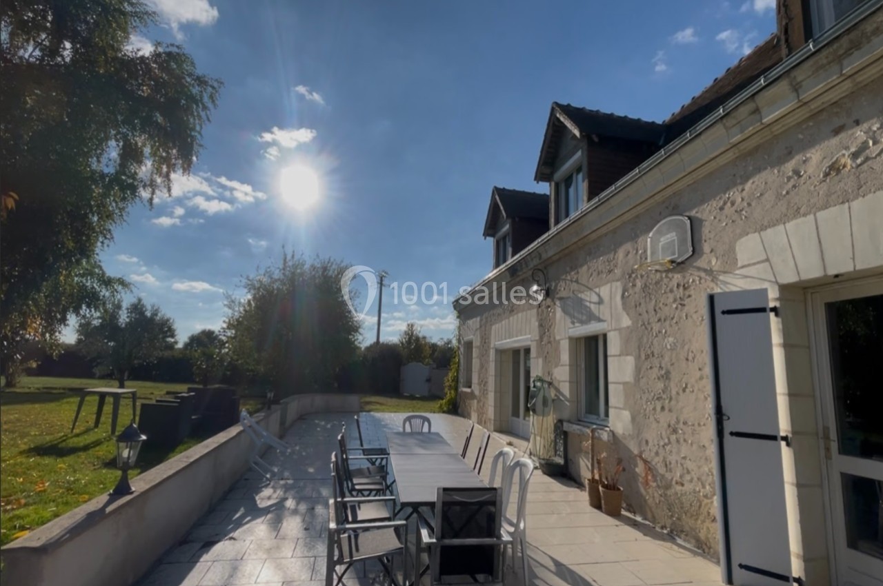 Terrasse ensoleillée avec une grande table et des chaises, adossée à une maison en pierre sous un ciel dégagé.