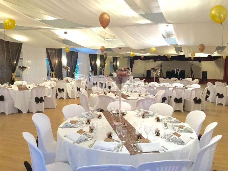 Salle de réception décorée avec des tables rondes dressées, nappes blanches et ballons suspendus au plafond.