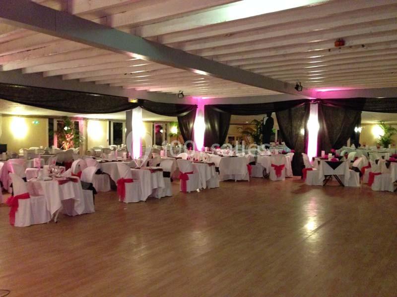 Salle de réception décorée avec des tables dressées, nappes blanches et nœuds rouges, éclairage rose et noir.