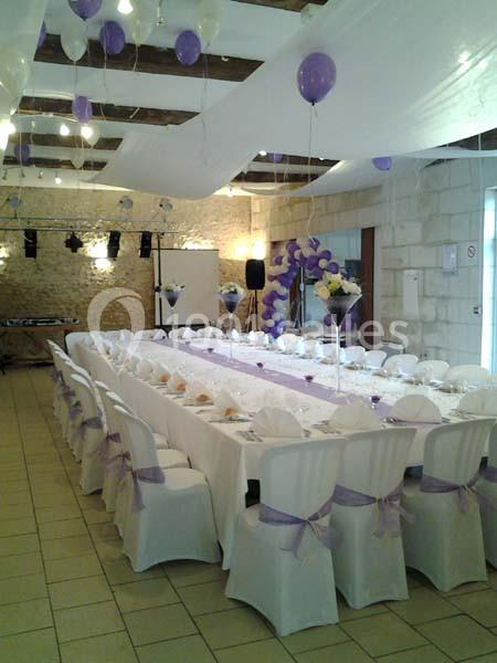 Salle décorée pour un événement, avec une grande table dressée, chaises blanches ornées de rubans violets et ballons…