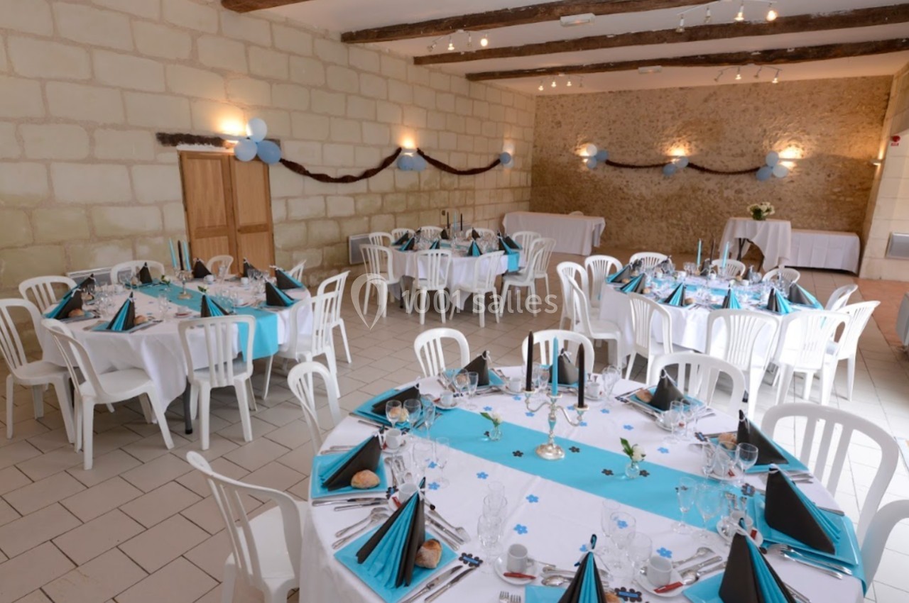 Salle décorée pour un événement avec tables rondes dressées, nappes blanches et bleues, chaises blanches et guirlandes…