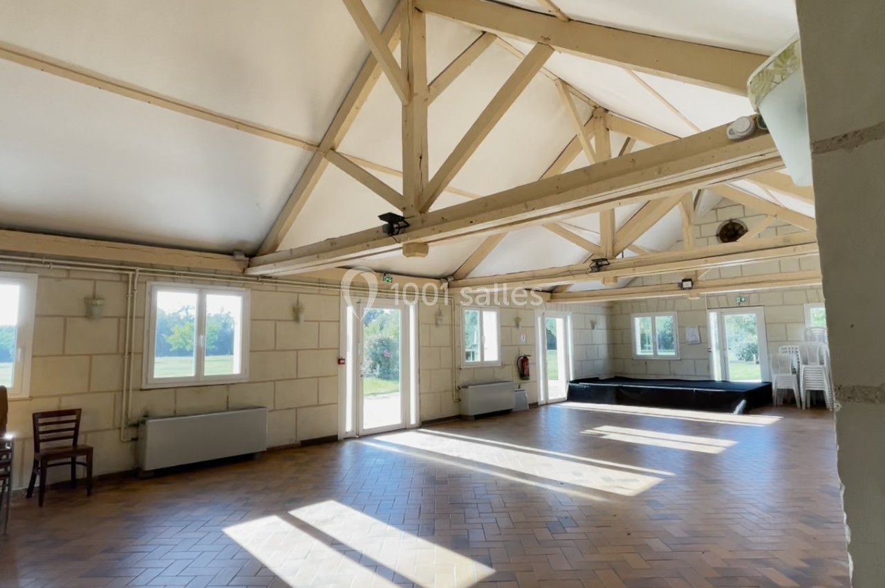 Salle lumineuse avec charpente apparente, parquet en bois, grandes fenêtres et empilement de chaises dans un coin.
