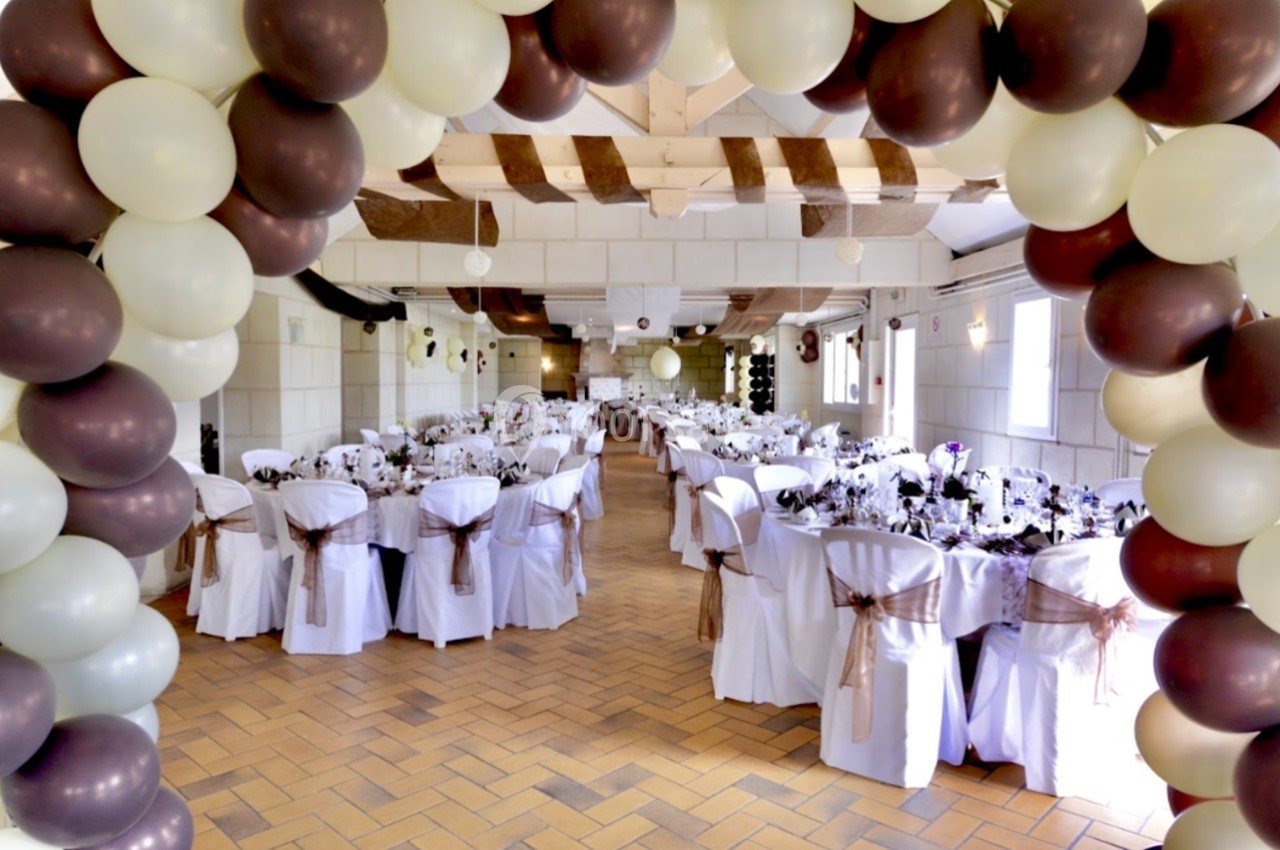 Salle de réception décorée pour un événement, avec des tables dressées et une arche de ballons en premier plan.