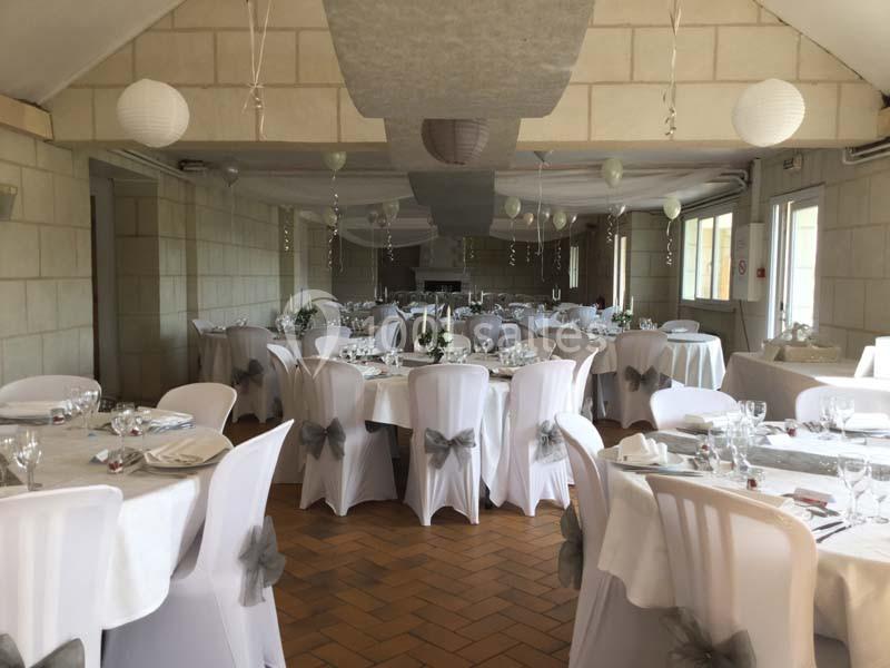 Salle de réception décorée avec des tables rondes, nappes blanches, chaises ornées de nœuds gris et lanternes suspendues.