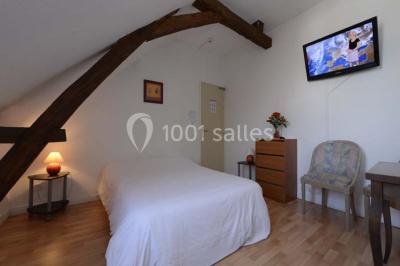 Chambre mansardée avec lit double, poutre apparente, télévision murale, commode, chaise et lampe de chevet.