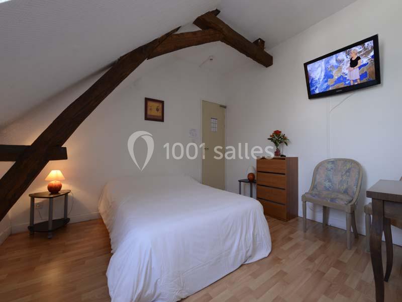 Chambre mansardée avec lit double, poutre apparente, télévision murale, commode, chaise et lampe de chevet.