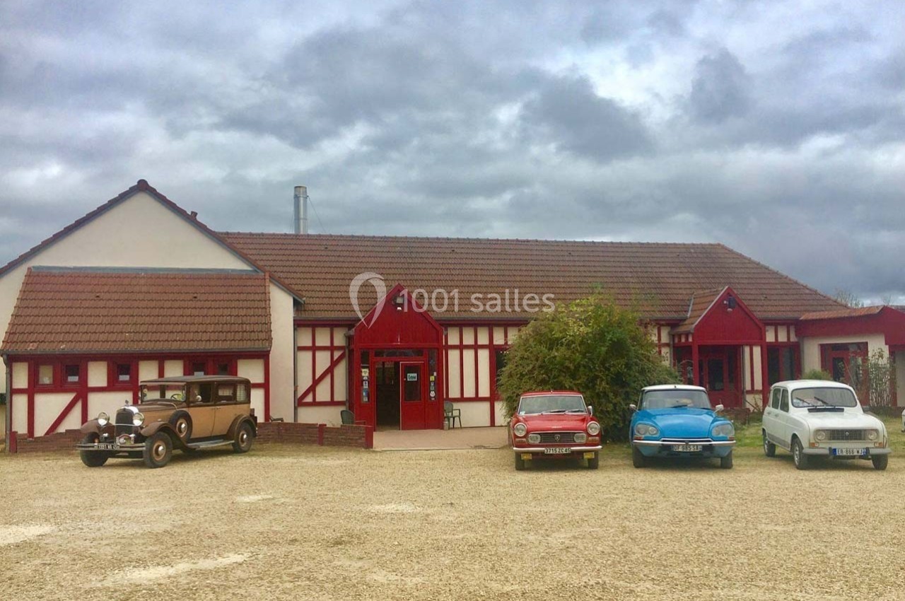 Façade d'un bâtiment à colombages rouges avec quatre voitures anciennes stationnées devant sur un sol gravillonné.